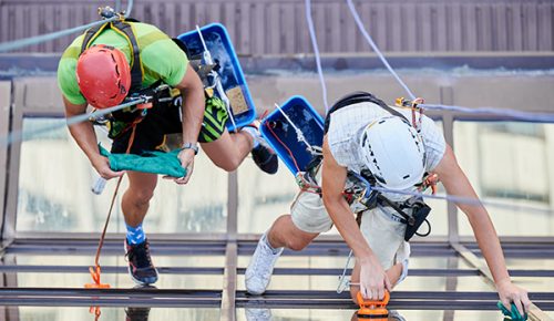 Industrial mountaineering. Top view of professional cleaners hanging on ropes while cleaning skyscraper windows. Two men window washers using cleaning tools while washing building facade.